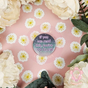 以下が含まれることがあります： 銀河のような背景に「If you can read this, you're TOO CLOSE」という紫色のテキストが書かれた丸いボタンです。