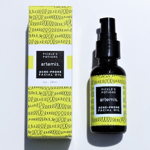 May include: A black glass bottle of "Pickle's Potions" Artemis acne-prone facial oil, next to a matching box. The box and bottle label have a yellow background with a squiggly pattern and white text. The bottle contains 1 fl oz (28 ml).