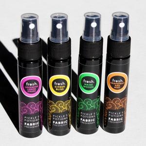 Puede incluir: Cuatro peque&ntilde;os frascos pulverizadores negros con etiquetas de diferentes colores. Las etiquetas tienen el texto "fresh." y una descripci&oacute;n del aroma. Los frascos est&aacute;n etiquetados con "Lavender Lemon Grass", "Citrus Bliss", "Maine Woods" y "Grapefruit and Mint". Los frascos est&aacute;n etiquetados con "Pickle's Potions Fabric Freshener".