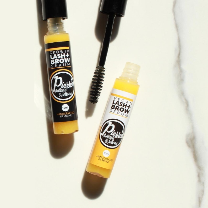 K&ouml;nnte beinhalten: Zwei gelb get&ouml;nte Wimpern- und Augenbrauenserumflaschen mit schwarzen Kappen und Applikatoren. Eine Flasche tr&auml;gt die Aufschrift "GROWTH LASH+ BROW SERUM" und die andere "VEGAN LASH+ BROW SERUM". Beide Flaschen haben das Logo "Pickle Potions & Lotions".