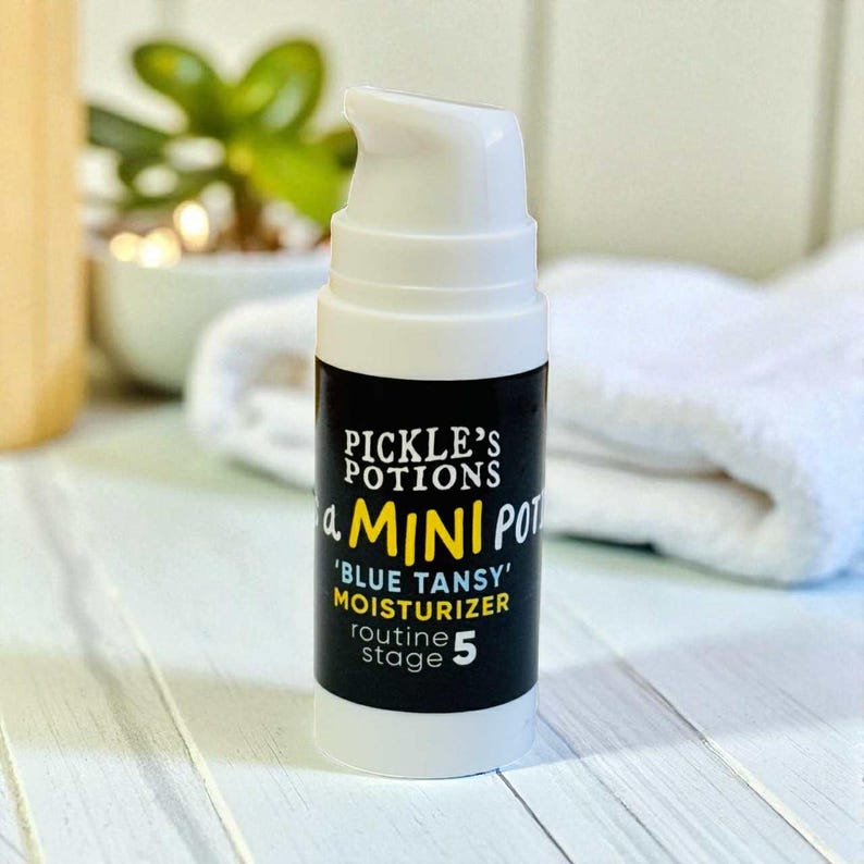May include: A white and black bottle of 'Blue Tansy' moisturizer from Pickle's Potions. The bottle has a white pump top and black label with white and yellow text. The text reads 'a MINI POT' and 'routine stage 5'.