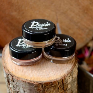 May include: Three small glass jars with black lids are stacked on a wooden surface. Each jar has a label that reads "Pickle's Potions & Lotions" in white script. The jars are filled with a light-colored product, and the background is a blurred wooden texture.