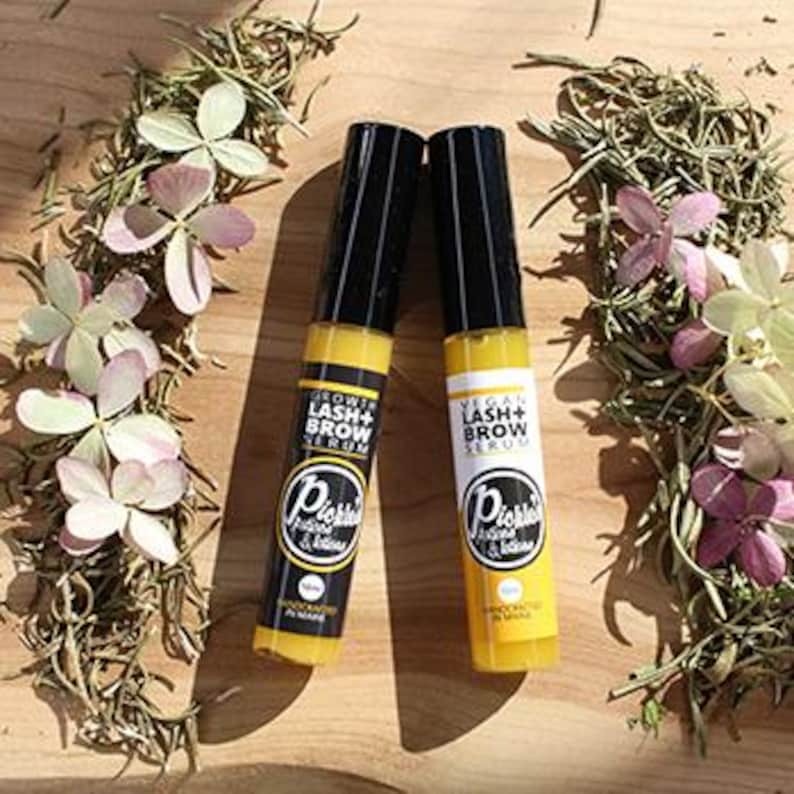 K&ouml;nnte beinhalten: Zwei gelb-schwarze Flaschen Wimpern- und Augenbrauenserum mit dem Text "Grow Lash + Brow Serum" und "Vital Lash + Brow Serum". Die Flaschen sind von Rosmarinzweigen und kleinen rosa und wei&szlig;en Bl&uuml;ten umgeben. Der Text "Pickles & Botanicals" ist ebenfalls sichtbar.