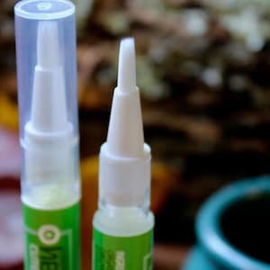 May include: Close-up of two white applicator pens with clear caps. The pens have a white brush tip and a green and white label. The background is blurred, with hints of green and brown.