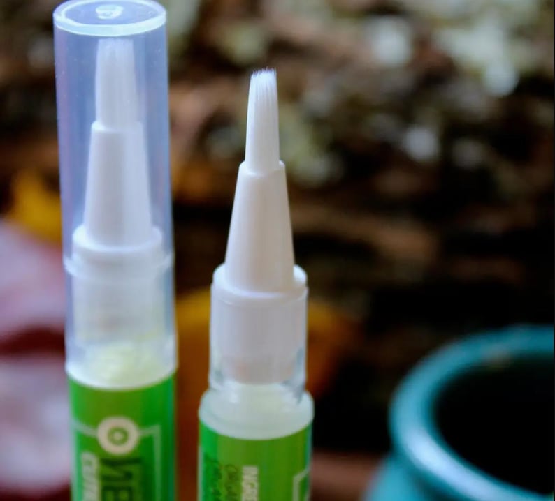 May include: Close-up of two white applicator pens with clear caps. The pens have a white brush tip and a green and white label. The background is blurred, with hints of green and brown.
