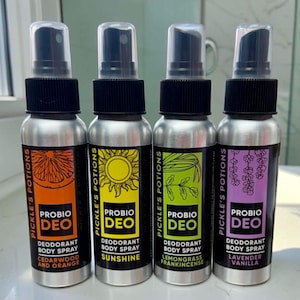 Op de afbeelding: Vier zilveren metalen spuitflessen met Probio Deo deodorant bodyspray van Pickle's Potions. De oranje, gele, groene en paarse etiketten bevatten illustraties van citrus, een zon, bladeren en lavendel, met de namen van de geuren eronder.