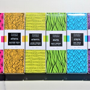 May include: A collection of six facial oil boxes with colorful designs and text. The boxes are labeled "Rosacea," "Athena," "Artemis," "Aura," "Sirena," and "Apollo." Each box has a unique pattern and color scheme, including pink, yellow, green, blue, and purple.