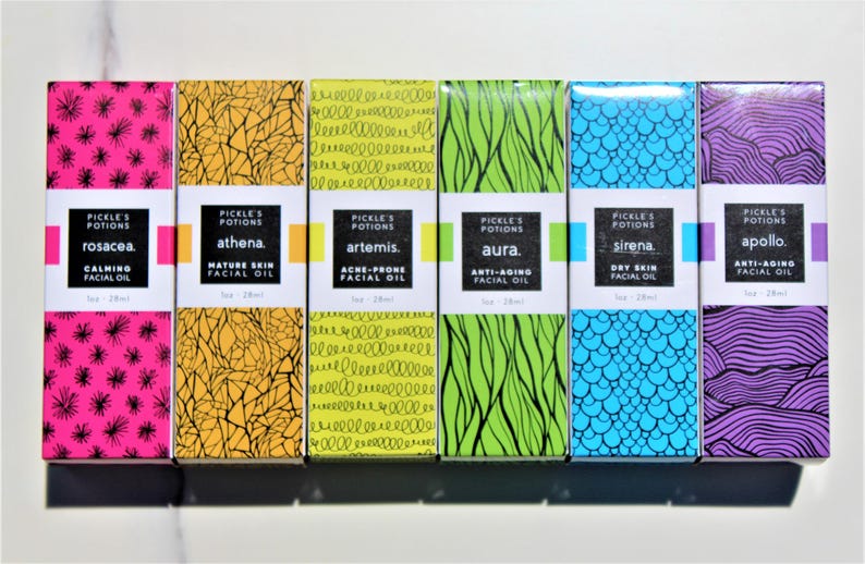 May include: A collection of six facial oil boxes with colorful designs and text. The boxes are labeled "Rosacea," "Athena," "Artemis," "Aura," "Sirena," and "Apollo." Each box has a unique pattern and color scheme, including pink, yellow, green, blue, and purple.