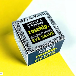Puede incluir: Una pequeña caja cuadrada de bálsamo para ojos iluminador de rosa mosqueta de Pickle's Potions. La caja es azul marino con texto blanco y negro y un patrón de remolino decorativo. El texto incluye "rosehip" en color verde brillante.