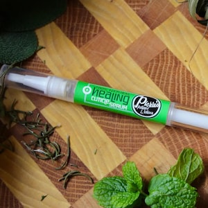 May include: A green and white cuticle serum pen with the text "Healing Cuticle Serum" and the brand name "Pickles Potions & Lotions". The pen is lying on a wooden surface with herbs and leaves.