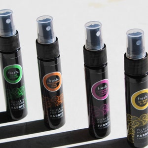 Puede incluir: Cuatro peque&ntilde;os frascos pulverizadores negros con etiquetas de diferentes colores. Las etiquetas tienen el texto "fresh." y una descripci&oacute;n del aroma. Los frascos est&aacute;n etiquetados como "Pickle's Potions Fabric Freshener".