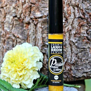 K&ouml;nnte beinhalten: Eine schwarze und gelbe Growth Lash + Brow Serum Flasche mit schwarzem Deckel. Auf der Flasche steht "Pickles Potions & Lotions" und "Handcrafted in Maine". Eine gelbe Blume und gr&uuml;ne Bl&auml;tter sind im Hintergrund.