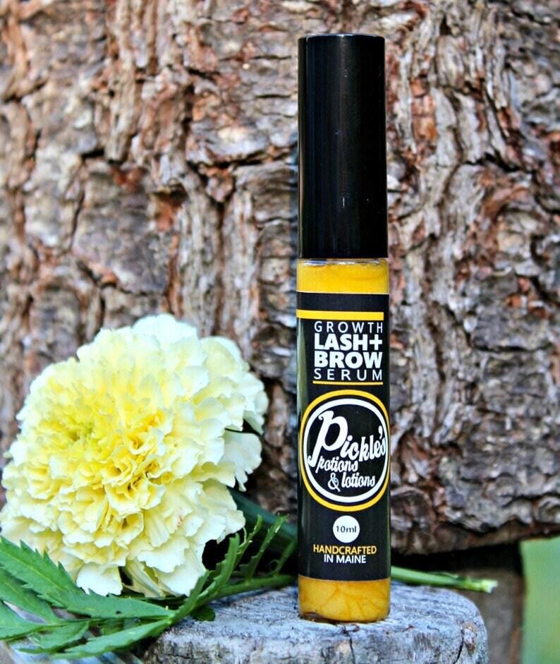 K&ouml;nnte beinhalten: Eine schwarze und gelbe Growth Lash + Brow Serum Flasche mit schwarzem Deckel. Auf der Flasche steht "Pickles Potions & Lotions" und "Handcrafted in Maine". Eine gelbe Blume und gr&uuml;ne Bl&auml;tter sind im Hintergrund.