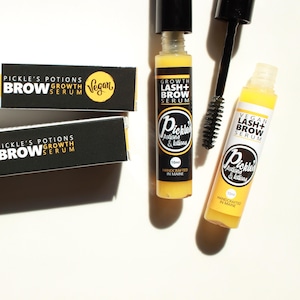 K&ouml;nnte beinhalten: Draufsicht auf Sch&ouml;nheitsprodukte, darunter zwei gelb-wei&szlig;e Flaschen Wimpern- und Augenbrauenserum, eine schwarze Mascarab&uuml;rste und zwei schwarze Schachteln. Die Flaschen und Schachteln tragen die Aufschrift "Pickle's Potions" und "Vegan Lash + Brow Serum."