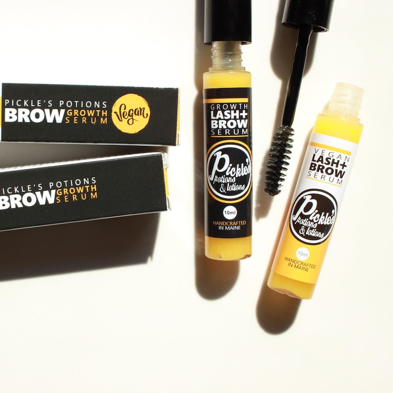 K&ouml;nnte beinhalten: Draufsicht auf Sch&ouml;nheitsprodukte, darunter zwei gelb-wei&szlig;e Flaschen Wimpern- und Augenbrauenserum, eine schwarze Mascarab&uuml;rste und zwei schwarze Schachteln. Die Flaschen und Schachteln tragen die Aufschrift "Pickle's Potions" und "Vegan Lash + Brow Serum."