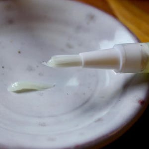 May include: A white pen-like applicator with a brush tip, dispensing a small amount of pale, creamy substance onto a white ceramic dish. The dish has a speckled, textured surface, and the applicator is angled towards the cream.