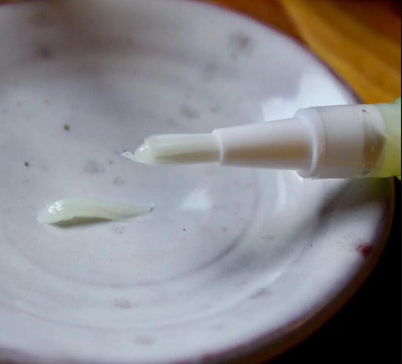 May include: A white pen-like applicator with a brush tip, dispensing a small amount of pale, creamy substance onto a white ceramic dish. The dish has a speckled, textured surface, and the applicator is angled towards the cream.