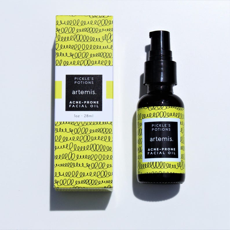 May include: A black glass bottle of "Pickle's Potions" Artemis acne-prone facial oil, next to a matching box. The box and bottle label have a yellow background with a squiggly pattern and white text. The bottle contains 1 fl oz (28 ml).