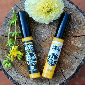 K&ouml;nnte beinhalten: Zwei gelb-schwarze Wimpern- und Augenbrauenserum-Flaschen mit dem Text "Growth Lash + Brow Serum" und "Vegan Lash + Brow Serum". Die Flaschen sind mit "Pickles Potions & Lotions" und "Handcrafted in Maine" beschriftet. Eine gelbe Blume und Gr&uuml;nzeug im Hintergrund.