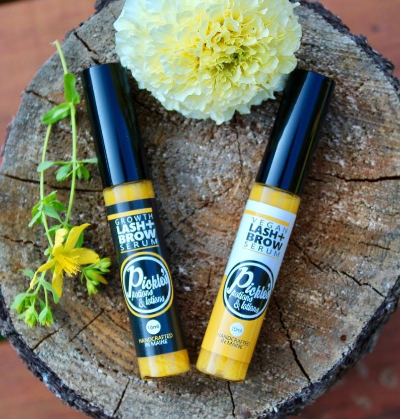 K&ouml;nnte beinhalten: Zwei gelb-schwarze Wimpern- und Augenbrauenserum-Flaschen mit dem Text "Growth Lash + Brow Serum" und "Vegan Lash + Brow Serum". Die Flaschen sind mit "Pickles Potions & Lotions" und "Handcrafted in Maine" beschriftet. Eine gelbe Blume und Gr&uuml;nzeug im Hintergrund.