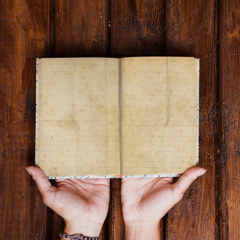 May include: An open notebook with lined pages, resting on a brown wooden surface. The notebook is held open by two hands.