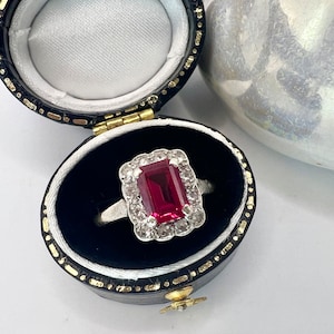May include: A red gemstone ring with a square cut stone surrounded by smaller diamonds set in a silver band. The ring is in a black velvet ring box.