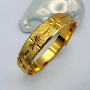 May include: A gold-toned hinged bangle bracelet featuring a textured surface with engraved starburst patterns. The bracelet has a polished interior and a secure clasp mechanism, showcasing a bright metallic finish throughout the entire piece.