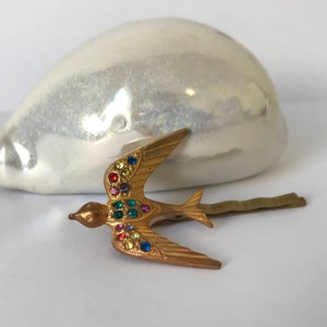May include: A gold-colored metal hair clip in the shape of a bird, adorned with colorful rhinestones. The bird is in flight, with its wings spread. The clip is a decorative accessory.