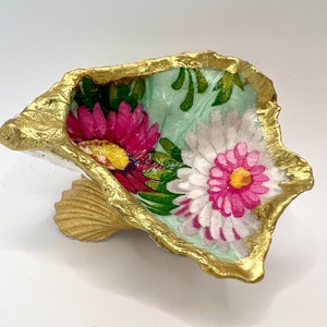 May include: A decorative oyster shell dish with a gold-painted rim and base. The interior features a light blue background with a floral design of pink and white daisies. The shell is a unique, handcrafted item.