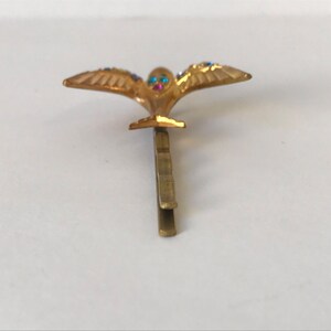 May include: A gold-colored hair clip featuring a stylized bird with outstretched wings. The bird's eyes are adorned with blue and pink gemstones, and the wings are embellished with blue gemstones. The clip is designed to secure hair.