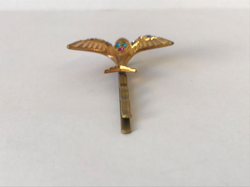 May include: A gold-colored hair clip featuring a stylized bird with outstretched wings. The bird's eyes are adorned with blue and pink gemstones, and the wings are embellished with blue gemstones. The clip is designed to secure hair.