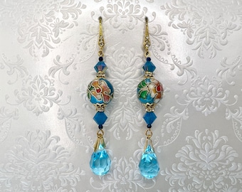 May include: Gold-tone dangle earrings with blue and floral accents. Each earring features a gold hook with clear crystals, a blue bead, a floral bead, and a large blue teardrop-shaped crystal. The floral beads have a light blue background with red, green, and white floral designs.