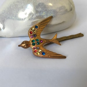 May include: A gold-toned metal hair clip in the shape of a bird with outstretched wings. The bird is adorned with colorful, sparkling gemstones. The clip is designed to secure hair and adds a decorative touch.