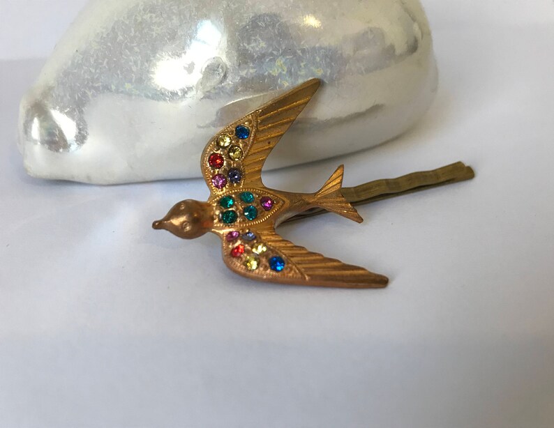 May include: A gold-toned metal hair clip in the shape of a bird with outstretched wings. The bird is adorned with colorful, sparkling gemstones. The clip is designed to secure hair and adds a decorative touch.