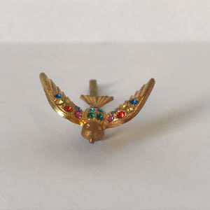 May include: A gold-colored bird-shaped brooch with outstretched wings. The wings are adorned with colorful, sparkling gemstones. The bird has a small, detailed head and a pin on the back for attachment. The brooch is set against a white background.