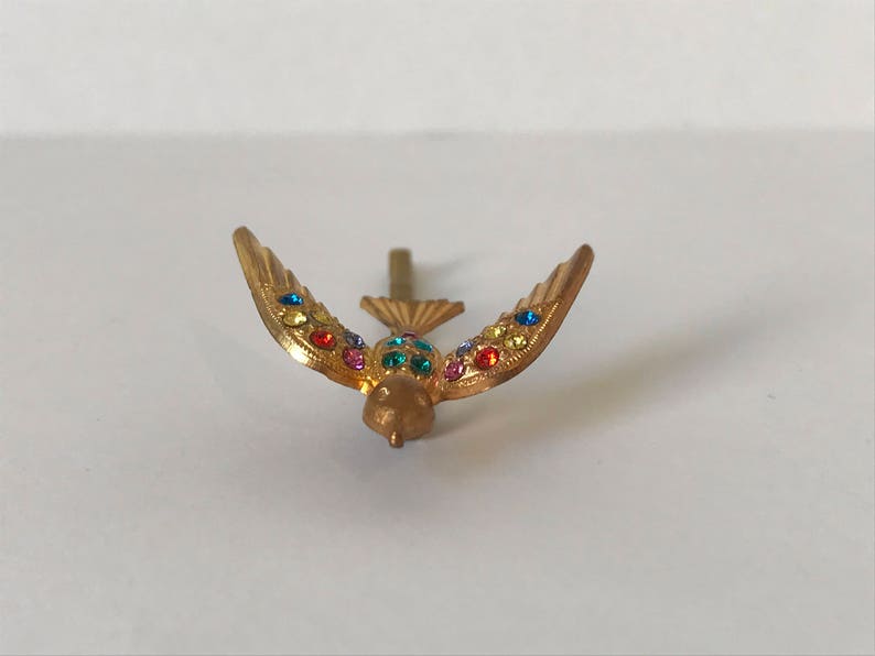 May include: A gold-colored bird-shaped brooch with outstretched wings. The wings are adorned with colorful, sparkling gemstones. The bird has a small, detailed head and a pin on the back for attachment. The brooch is set against a white background.