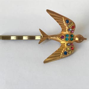 May include: Gold-toned metal hair barrette featuring a stylized bird in flight. The bird is adorned with colorful rhinestones in shades of red, blue, yellow, and green. The barrette is a classic accessory.