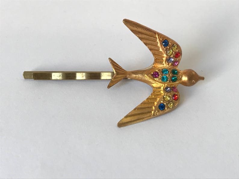 May include: Gold-toned metal hair barrette featuring a stylized bird in flight. The bird is adorned with colorful rhinestones in shades of red, blue, yellow, and green. The barrette is a classic accessory.