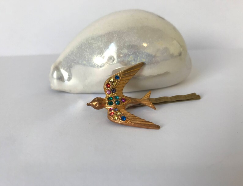 May include: A gold-colored metal hair clip in the shape of a bird, adorned with colorful rhinestones. The bird is in flight, with its wings spread. The clip is a decorative accessory.