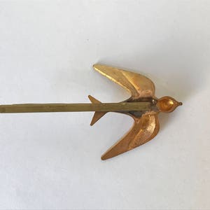 May include: A gold-colored metal hair barrette in the shape of a swallow bird. The bird has outstretched wings and a long, straight pin for securing it in the hair. The barrette is a decorative hair accessory.