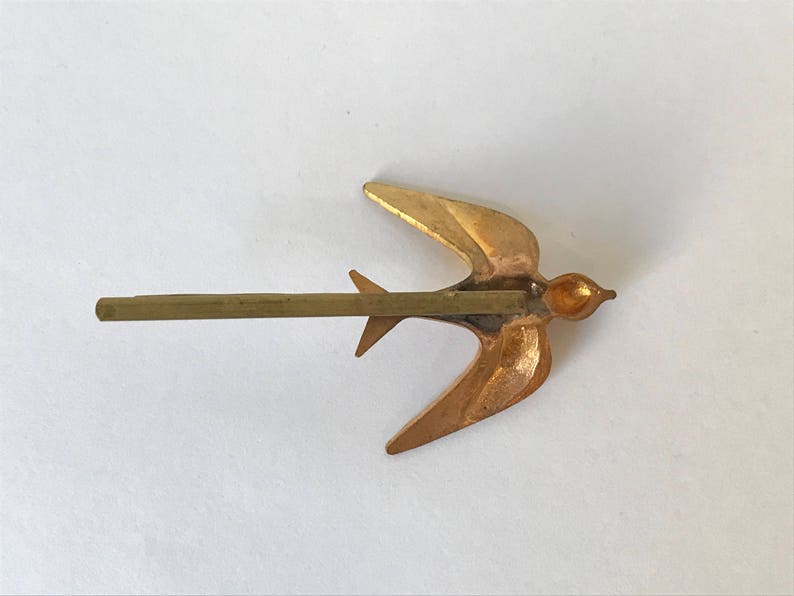 May include: A gold-colored metal hair barrette in the shape of a swallow bird. The bird has outstretched wings and a long, straight pin for securing it in the hair. The barrette is a decorative hair accessory.