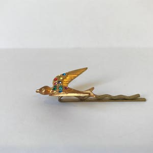 May include: A gold-toned hair bobby pin featuring a stylized bird in flight. The bird's wing is adorned with colorful rhinestones. The pin has a simple, wavy design.