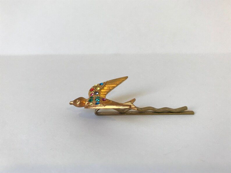 May include: A gold-toned hair bobby pin featuring a stylized bird in flight. The bird's wing is adorned with colorful rhinestones. The pin has a simple, wavy design.