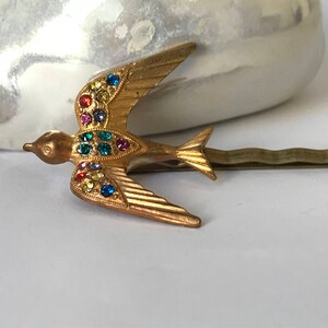 May include: Gold-toned metal bird hair clip with colorful rhinestones. The bird is in flight, with wings spread and tail feathers detailed. The clip is a bobby pin style, suitable for securing hair. The bird's body is adorned with various colored rhinestones.