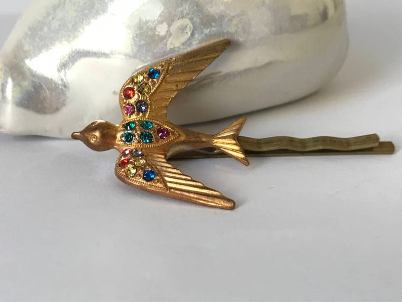 May include: Gold-toned metal bird hair clip with colorful rhinestones. The bird is in flight, with wings spread and tail feathers detailed. The clip is a bobby pin style, suitable for securing hair. The bird's body is adorned with various colored rhinestones.