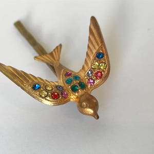 May include: A gold-colored metal bird-shaped hair clip with outstretched wings. The bird is adorned with colorful rhinestones in shades of blue, yellow, green, and red. The clip has a simple, straight pin.