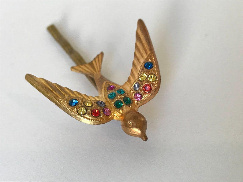 May include: A gold-colored metal bird-shaped hair clip with outstretched wings. The bird is adorned with colorful rhinestones in shades of blue, yellow, green, and red. The clip has a simple, straight pin.