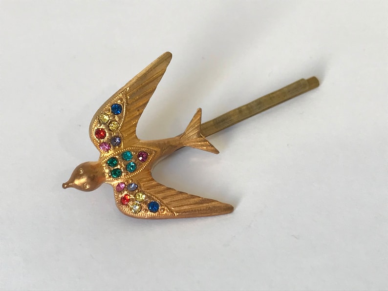 May include: A gold-toned metal hair clip in the shape of a bird. The bird's wings and body are adorned with colorful rhinestones. The clip has a simple, straight pin design.
