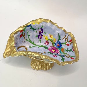 May include: A decorative oyster shell dish with a gold-painted rim and base. The interior features a light blue background with a floral pattern in various colors, including pink, yellow, and purple. The dish is designed to hold small items.