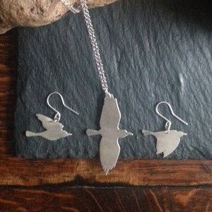 Puede incluir: Un conjunto de collar y pendientes de plata con un diseño de silueta de pájaro. El collar tiene una cadena delicada y los pendientes son de estilo colgante.